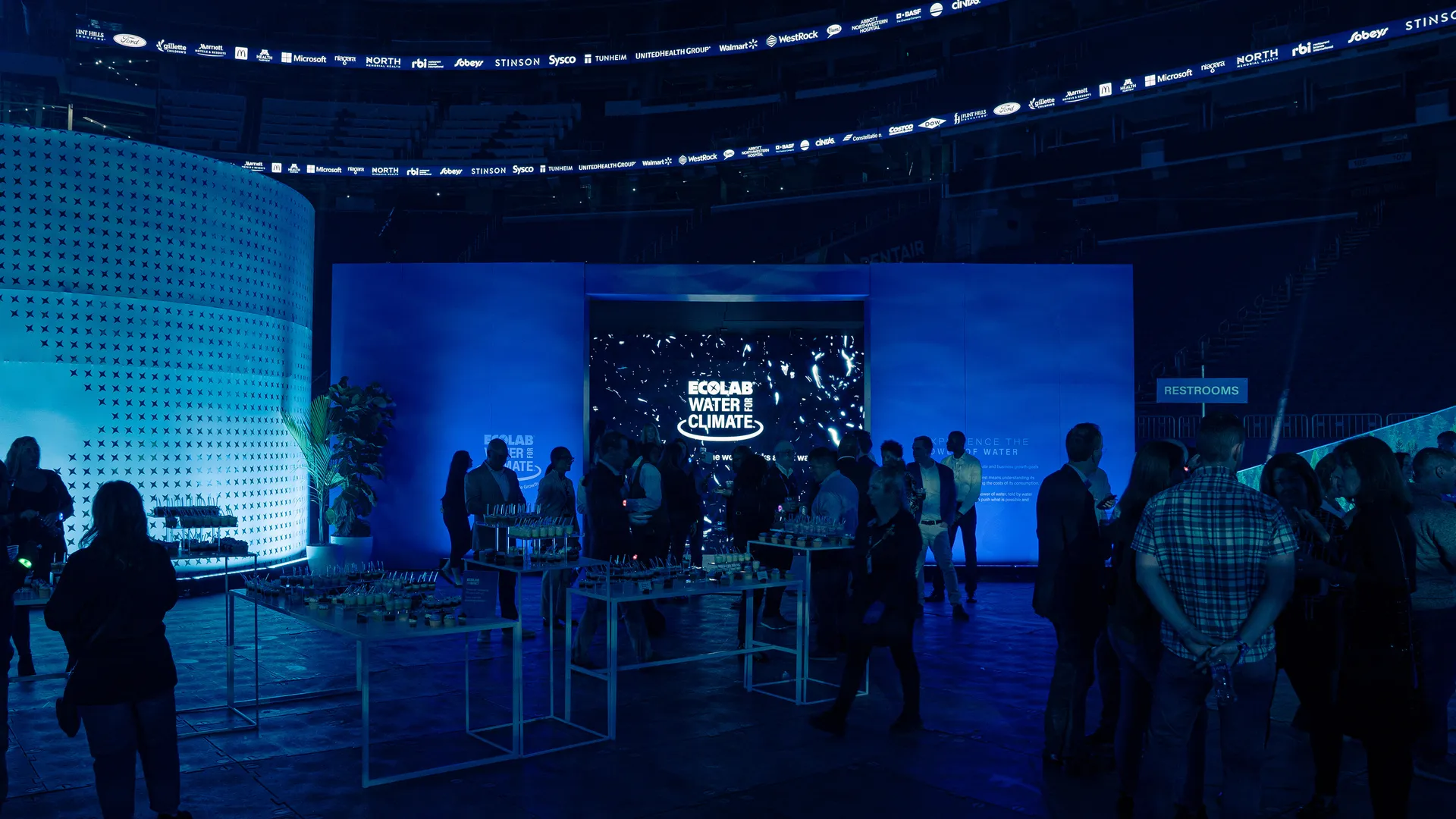 A photo of the Power of Water installation at the US Bank Stadium from outside lit with blue lighting. The Ecolab Water for Climate logo prominently displayed on the screen. many people standing around enjoying drinks and delicious treats.